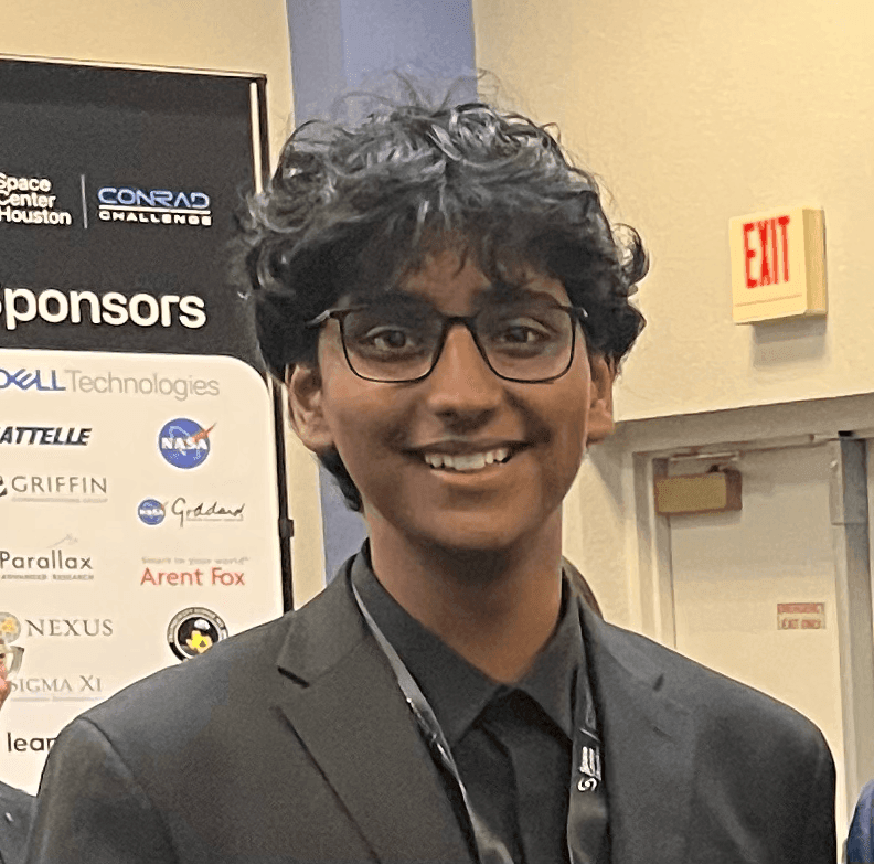 Person in a suit at an event with various company logos on a sponsor board behind them.