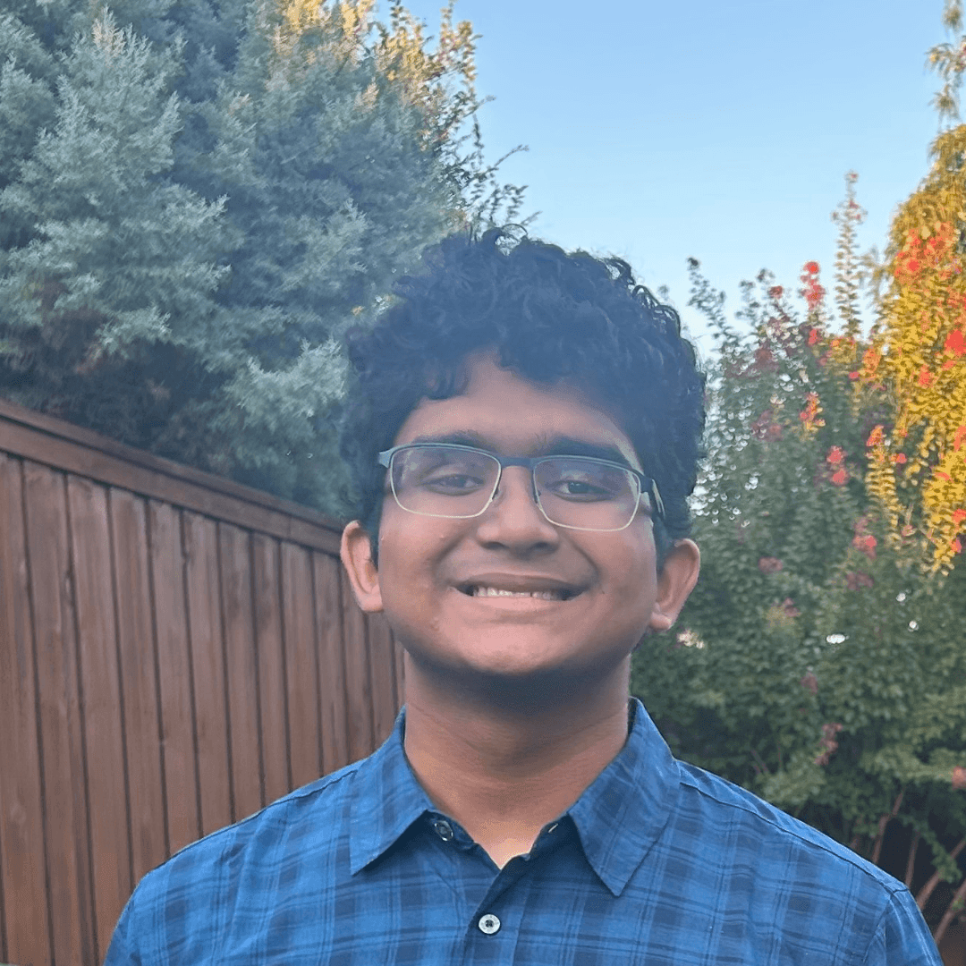 Person with glasses smiling outdoors, standing in front of a wooden fence and trees.